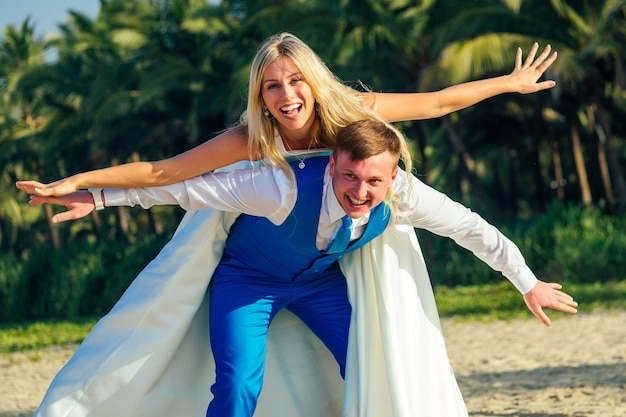 Przystojny pan młody w eleganckim garniturze i piękna panna młoda w sukni ślubnej bawią się i krzywią na plaży. koncepcja eleganckiej i bogatej ceremonii ślubnej na plaży.