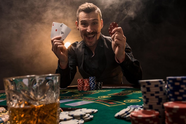 Przystojny pokerzysta z dwoma asami w rękach i żetonami, siedzący przy stole pokerowym w ciemnym pokoju pełnym dymu papierosowego.
