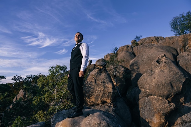 Przystojny poważny mężczyzna w garniturze stoi na szczycie skał na tle błękitnego nieba i patrzy w dal