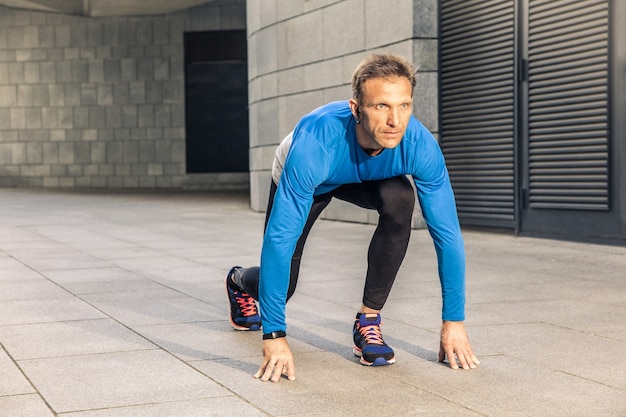 Przystojny poważny mężczyzna w średnim wieku w niebieskim mundurze sportowym i słuchawkach i monitorze fitness działa w mieście.