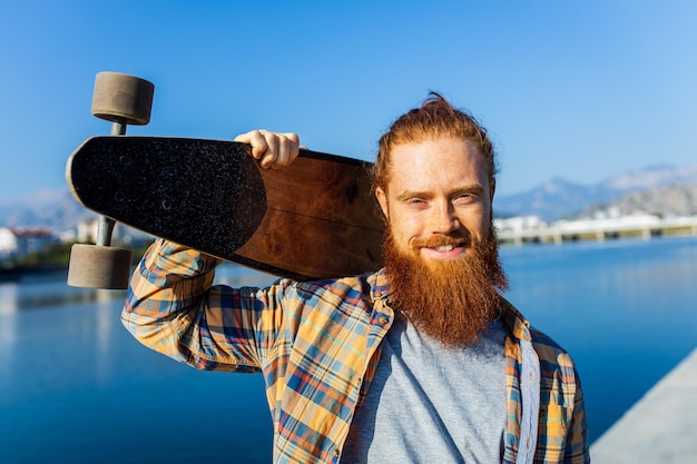 Przystojny rudowłosy mężczyzna z długą brodą na deskorolce w pobliżu rzeki w słoneczny letni dzień