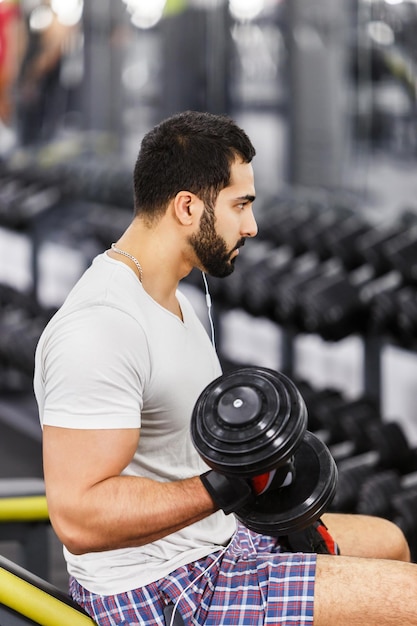 Przystojny seksowny brodaty mężczyzna nosi białą koszulkę i słuchawki ćwiczy biceps z hantlami na siłowni