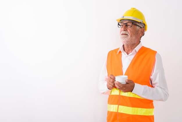 przystojny starszy brodaty mężczyzna pracownik budowlany myśli i trzyma filiżankę kawy podczas noszenia okularów na białym tle