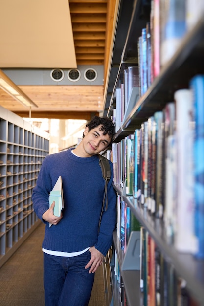 Przystojny student uśmiecha się patrząc na kamerę odwiedzając bibliotekę uniwersytecką w celu przygotowania do egzaminu