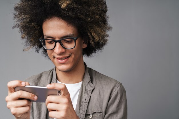 przystojny szczęśliwy mężczyzna z afro fryzurą uśmiechający się i grający w grę wideo na telefonie komórkowym odizolowanym na szaro