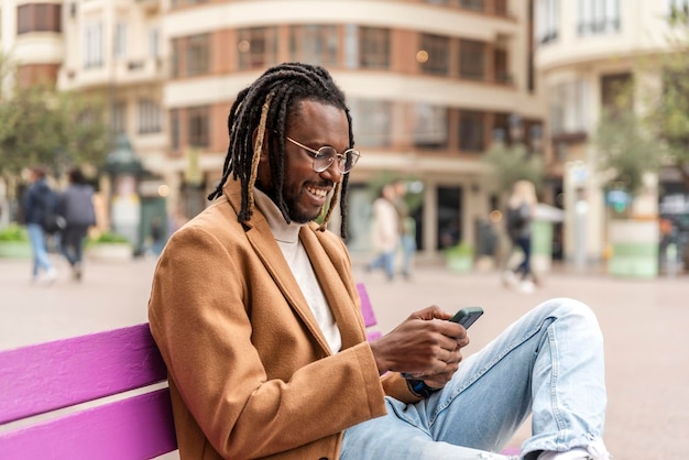 Przystojny uśmiechnięty mężczyzna siedzi na ławce w mieście i używa swojego telefonu