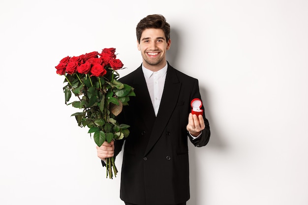 Przystojny Uśmiechnięty Mężczyzna W Czarnym Garniturze, Trzymając Róże I Pierścionek Zaręczynowy