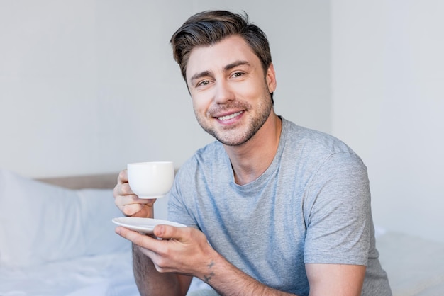 Przystojny uśmiechnięty mężczyzna w szarej koszulce trzymający filiżankę kawy i patrzący na kamerę