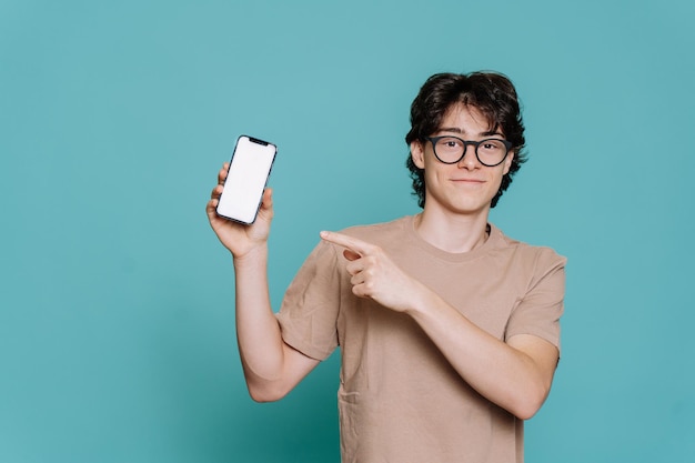 Przystojny włoski uczeń w okularach i beżowej koszulce odizolowany na turkusowym tle studia, wskazujący na telefon z pustym ekranem patrzy na aparat Wesoły nastolatek trzymający smartfon Makieta Model