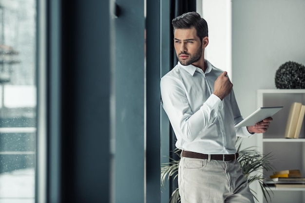 Przystojny zamyślony biznesmen trzyma cyfrowy tablet patrząc przez okno i myśląc stojąc w biurze