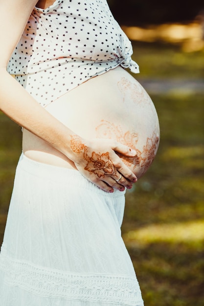 Przyszła mama w białej sukni trzyma nagi brzuszek pomalowany mehendi. Zewnętrzne zdjęcie brzucha kobiety w ciąży pomalowane mhendi