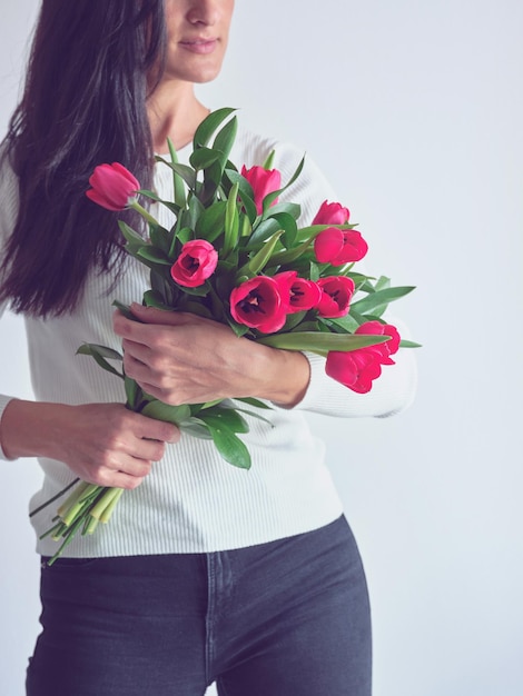 Przytnij anonimową kobietę, dotykając i wąchając bukiet jasnych tulipanów z przyjemnym uśmiechem na twarzy, stojąc w pobliżu białego tła