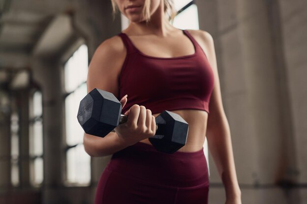 Przytnij nierozpoznawalną kobietę w stroju fitness, wykonując uginanie bicepsa z hantlami podczas treningu funkcjonalnego na siłowni