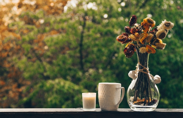 Przytulna jesienna martwa natura: filiżanka gorącej kawy z jesiennym bukietem kwiatów na parapecie vintage z szyszkami i deszczem na zewnątrz. Jesień. Apartament. Deszcz
