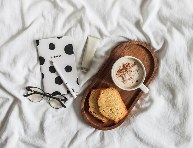 Przytulne poranne śniadanie w łóżku Kawa ciasto plasterki na drewnianej tacy planner notatnik krem do rąk na białych arkuszach widok z góry