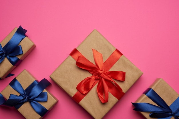Pudełka na prezenty zawinięte w brązowy papier i przewiązane czerwono-niebieską kokardką, prezenty na różowym tle, miejsce na tekst