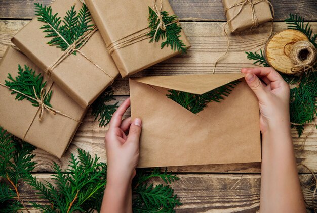 Pudełka zapakowane w papier pakowy, zielone gałązki na drewnianym tle. Kobiece ręce trzymają kopertę.