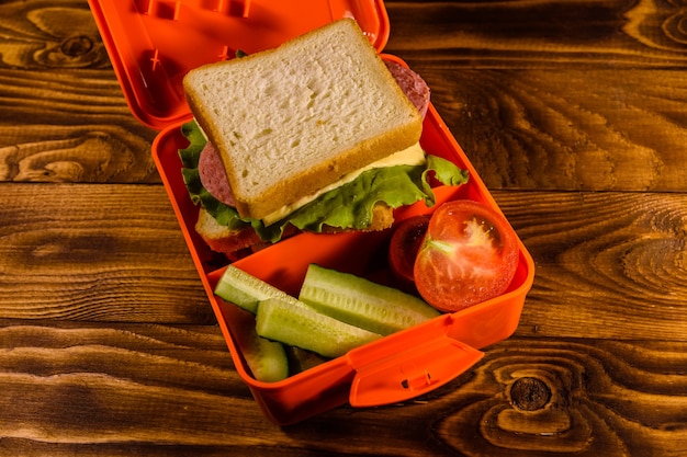 Pudełko na lunch z kanapką, ogórkami i pomidorami na rustykalnym drewnianym stole