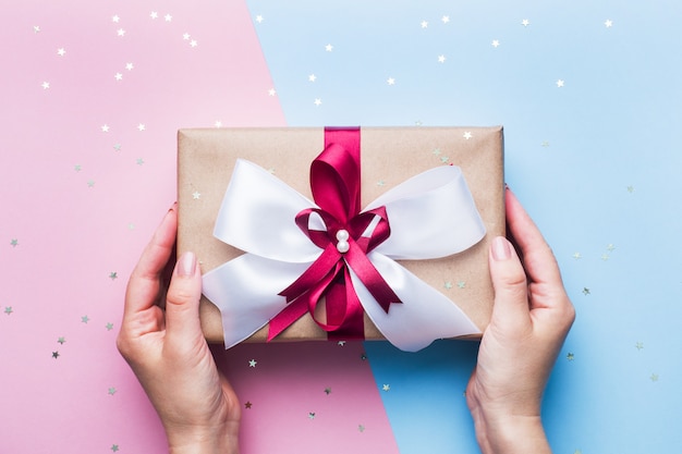 Pudełko prezentowe lub prezentowe z dużą kokardką w rękach kobiety na różowym niebieskim stole. Kompozycja płasko świecka na Boże Narodzenie, urodziny, dzień matki lub ślub.
