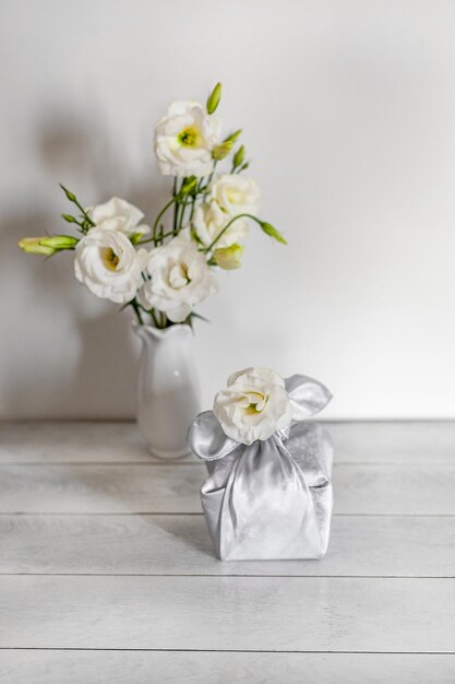 Pudełko prezentowe owinięte jedwabną tkaniną w technice Furoshiki białe kwiaty Eustoma lub Lisianthus w wazonie na jasnym drewnianym tle