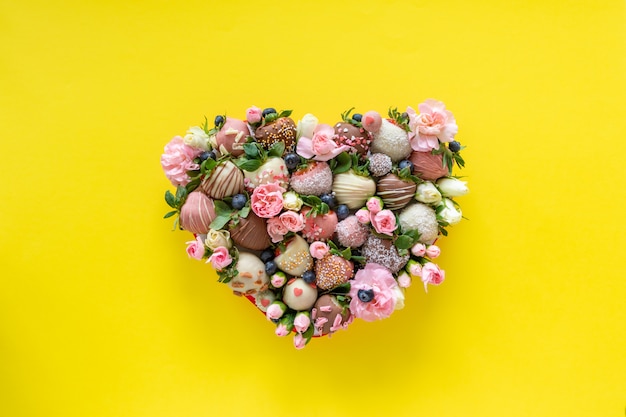Pudełko w kształcie serca z ręcznie robionymi czekoladowymi truskawkami z różnymi dodatkami i kwiatami jako prezent na Walentynki na żółtym tle