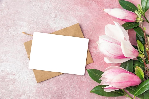 Pusta kartka z życzeniami z różowymi kwiatami magnolii i zielonymi liśćmi na różowym betonowym tle Płaska świecka makieta