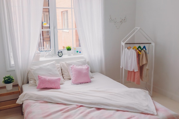 Pusta sypialnia z starannie zasłanym łóżkiem duże okno z białymi zasłonami ubrania na wieszakach Budzik na parapecie Przestronny przytulny pokój