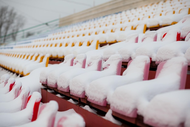 Puste siedzenia stadionu piłkarskiego na świeżym powietrzu pokryte śniegiem w zimie, lekkie opady śniegu. Plastikowe siedzenia w rzędzie na stadionie pokryte są śniegiem.