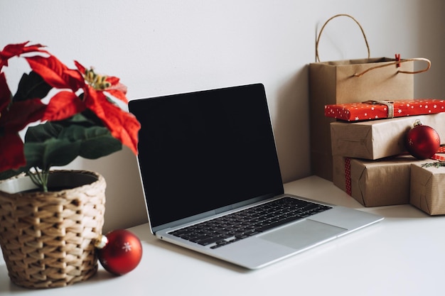 Pusty ekran laptopa z bożonarodzeniowymi kwiatami poinsecji i pudełkami prezentów na białym stole