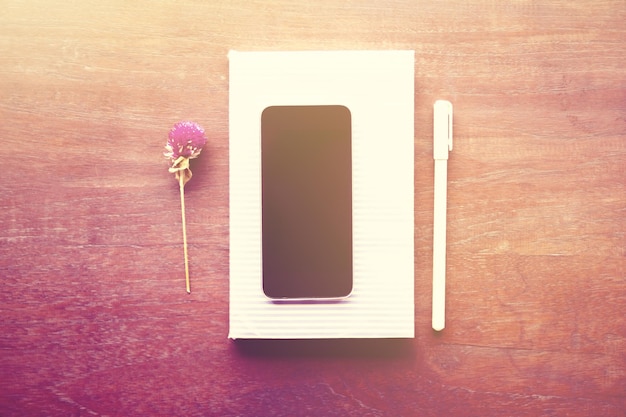 Pusty ekran smartfona na pustym pamiętniku z kwiatem i piórem na drewnianym stole.
