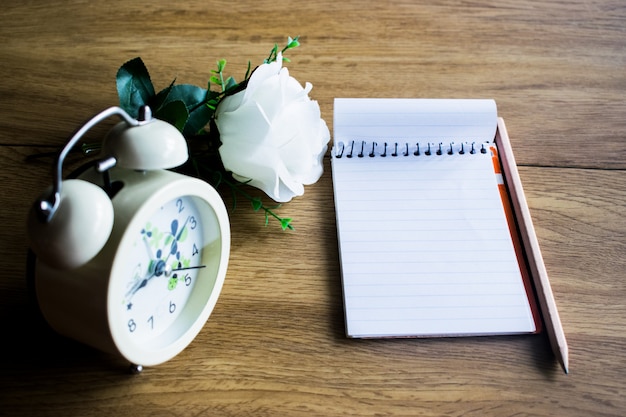 Pusty notatnik, ołówek, budzik i biała róża na drewnianym stole