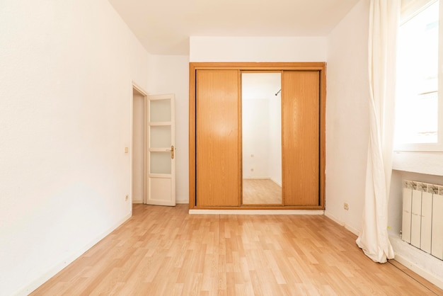 Pusty pokój z wbudowaną szafą z przesuwanymi lustrzanymi drzwiami w połączeniu z drewnem i białymi drewnianymi drzwiami oraz laminowaną podłogą