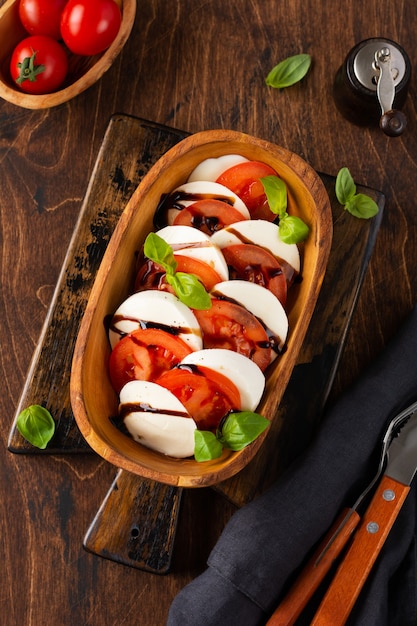 Pyszna włoska sałatka caprese z dojrzałymi pomidorami, świeżą bazylią ogrodową i serem mozzarella w drewnianej misce z oliwek na starym rustykalnym stole Widok z góry płaska leżanka