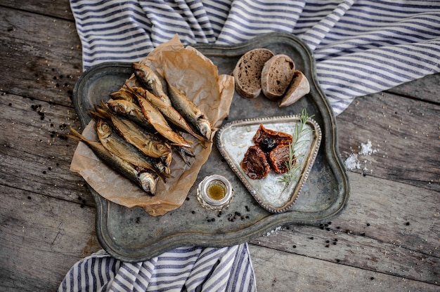 Pyszna złocista wędzona ryba ostroboka na papierze na metalowej tacy z suszonymi na słońcu pomidorami, oliwą i chlebem na szarej pasiastej serwetce na drewnianym stole.