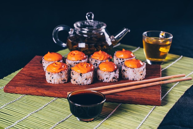 Pyszne bułki na drewnianej desce z pałeczkami i zieloną herbatą w czajniczku na dekoracyjnej bambusowej serwetce Zbliżenie smacznych sushi rolek na desce do krojenia na czarnym tle