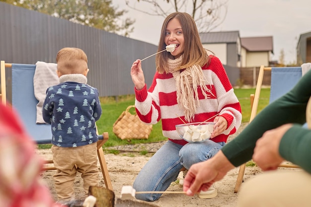 Pyszne Pianki. Młoda ładna Kobieta Z Długimi Blond Włosami W Pasiastym Swetrze Kucając Trzymając Talerz Odgryzający Pianki Patrząc Na Bok I Mąż Z Dziećmi Przy Ognisku Na Zewnątrz