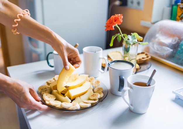 Pyszne pożywne śniadanie z kawą i herbatą, pokrojone na kawałki melon i banan, świeże tosty z masłem i orzechami czekają na stole czekając na towarzystwo lub rodzinę w przytulnej domowej kuchni rano