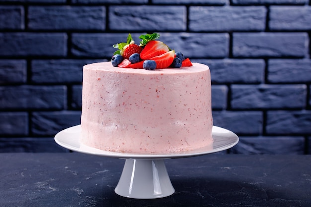 Pyszny i słodki cały różowy tort truskawkowy ozdobiony świeżymi owocami i listkami mięty na białej paterce z ceglaną ścianą