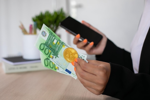 Rachunki euro i bitcoin w rękach kobiety z telefonem w miejscu pracy
