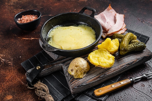 Zdjęcie raclette szwajcarski topiony ser z gotowanymi ziemniakami i szynką na drewnianej desce. ciemne tło. widok z góry.
