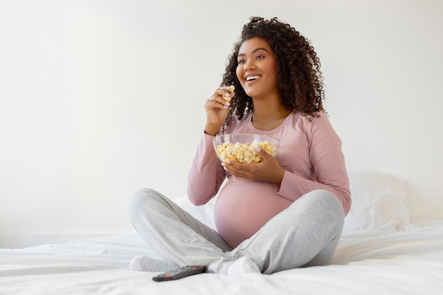 Radość w ciąży czarna kobieta jedząca popcorn siedząc w łóżku w domu