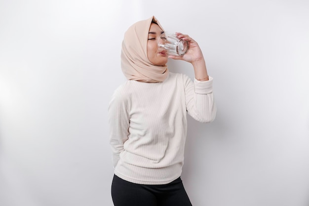 Radosna azjatycka muzułmanka w chustce na głowę pije szklankę wody na białym tle