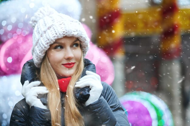 Radosna blond kobieta nosi czapkę z dzianiny spaceru na zimowych targach podczas opadów śniegu. Puste miejsce na tekst