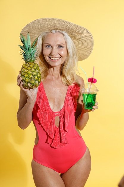 Radosna blondynka w czerwonym stroju kąpielowym i słomkowym kapeluszu uśmiecha się do kamery trzymającej świeże owoce ananasa