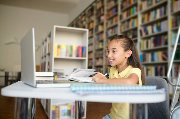 Radosna ciemnowłosa uczennica śni na jawie przy bibliotecznym stole