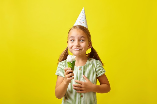Radosna dziewczynka w urodzinowym kapeluszu ma zaskoczoną minę, jedząc lody stoi na żółtym odosobnionym tle