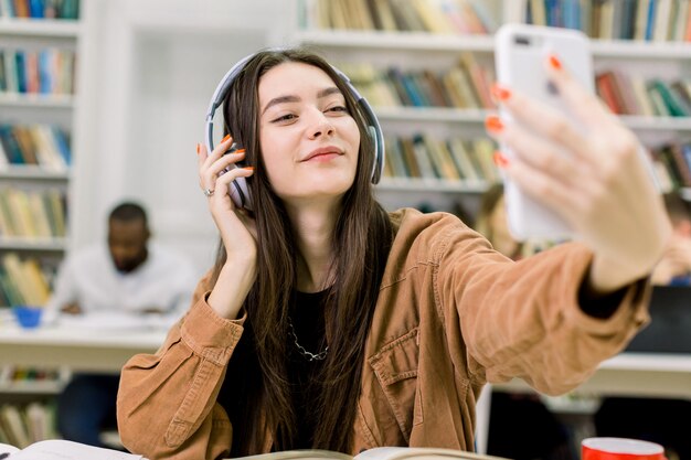 Radosna ładna młoda studentka słuchająca muzyki w słuchawkach i trzymająca inteligentny telefon, siedząc w nowoczesnej bibliotece i studiując ze swoimi wieloetnicznymi kolegami z grupy