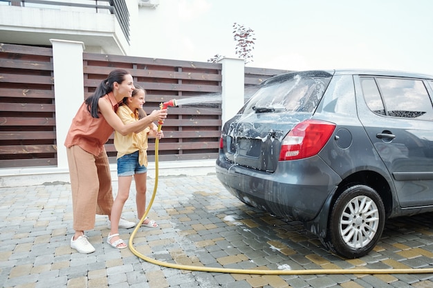 Radosna mama i córka spędzają czas razem myjąc samochód wodą za pomocą węża