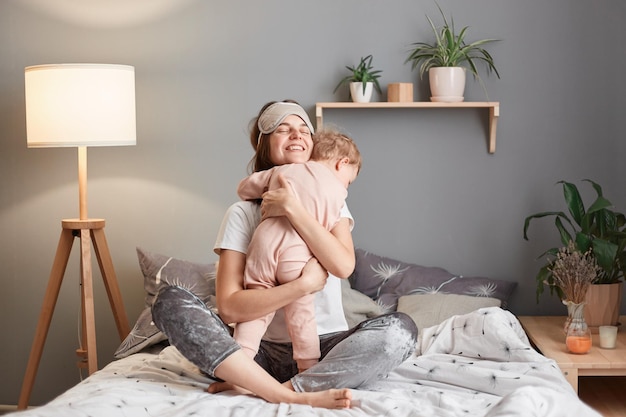 Radosna matka w masce do spania na czole przytulająca swoją córeczkę bawiącą się w łóżku u kobiety obejmującej dziecko, która chętnie spędza poranek razem z rodziną bawiąc się po przebudzeniu