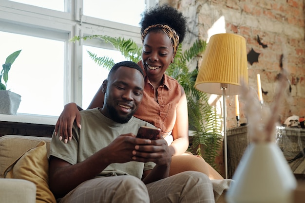 Radosna młoda afrykańska para w casualwear, patrząca na ekran smartfona trzymanego przez uśmiechniętego mężczyznę, siedząc przed kamerą przed oknem